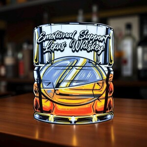 May include: A whiskey glass with the text "Emotional Support Lexus Whiskey" in a stylized font. The glass features an illustration of a car in shades of blue, orange, and yellow. The background is blurred, suggesting a bar setting.