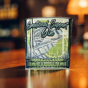 May include: A glass coaster with a graphic of a cocktail glass filled with ice, a lime wedge, and a blue and white space ship. The text "Emotional Support Gin" is printed on the coaster.