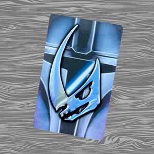 May include: A blue and white Mandalorian symbol pin with a textured finish. The pin is on a background of a blue and grey Mandalorian helmet.
