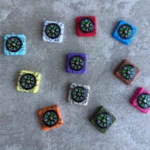 May include: A selection of square charms, each with a compass design. The compasses have black faces with green and white markings, set within colourful squares of blue, red, purple, brown, pink, yellow, orange, green, and white.