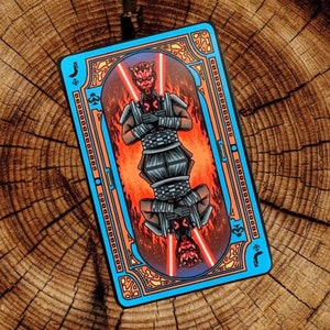 Pode incluir: Um baralho de cartas com fundo azul e moldura dourada. O cartão apresenta uma ilustração vermelha e preta de Darth Maul, um personagem da franquia Star Wars. O cartão é um Valete de Espadas.