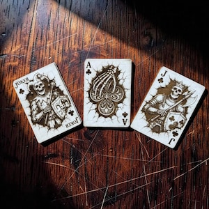 May include: Three playing cards with a gothic theme are displayed on a wooden surface. The cards feature skeletal figures and intricate designs. The cards are the Joker, Ace of Clubs, and Jack of Clubs.