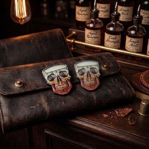 May include: A dark brown distressed leather wallet featuring two metallic skull-shaped embellishments on the front flap. The wallet is secured with two brass-colored snap buttons and has a textured, weathered appearance with visible grain details.