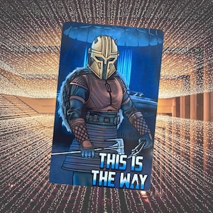 May include: A blue and gold illustration of a Mandalorian warrior with a helmet and armour. The text "This is the Way" is printed below the warrior.