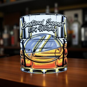 May include: A sticker featuring a stylized illustration of a whiskey glass. The glass is filled with amber liquid and ice, with the text "Emotional Support Ex Whiskey" in a script font. The design has a cartoonish style with bold outlines and bright colors.