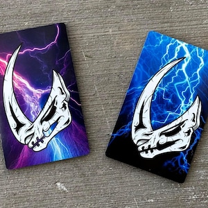 May include: Two rectangular cards with a white skull-like design on a black background. The cards have a blue and purple lightning bolt pattern.