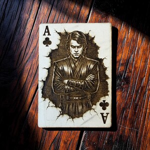 May include: Ace of Clubs playing card featuring a detailed illustration of a person with crossed arms. The card has a distressed, aged appearance with the suit symbols and the letter "A" visible. The card is set against a dark wooden surface.