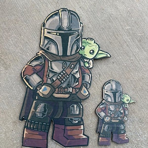 Puede incluir: Una figurita de dibujos animados de madera marrón y gris de un Mandaloriano con un personaje alienígena verde en su hombro. El Mandaloriano lleva un casco y una armadura, y el alienígena sostiene un objeto pequeño. La figurita está hecha de madera y tiene un acabado desgastado.
