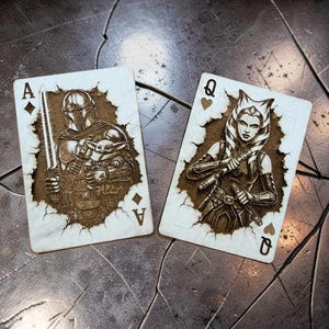 May include: Two playing cards featuring Star Wars characters. The Ace of Diamonds card depicts The Mandalorian with Grogu, and the Queen of Diamonds shows Ahsoka Tano. The cards have a detailed, engraved design with a brown and white color scheme.