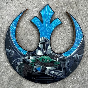 May include: A wooden cutout of the Rebel Alliance symbol with a blue and black illustration of the Mandalorian and Grogu inside. The Mandalorian is wearing his helmet and holding Grogu in his arms. The background is a blue and black nebula.