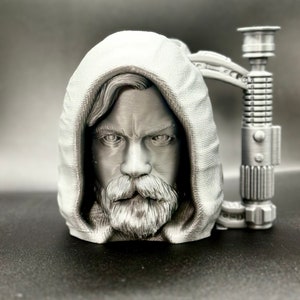 May include: A silver 3D printed mug featuring a detailed portrait of Luke Skywalker from Star Wars. The mug's handle is shaped like a lightsaber.