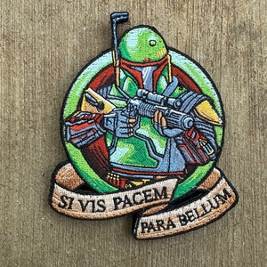 May include: A green and brown embroidered patch featuring a Star Wars character, Boba Fett, holding a blaster rifle. The patch has a green circle around the character and a brown banner below with the text "Si Vis Pacem Para Bellum".