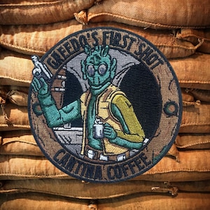Può includere: Toppa ricamata con un personaggio alieno verde che tiene una tazza di caffè. La toppa è circolare e ha il testo "Greedo's First Shot Cantina Coffee" attorno al bordo.