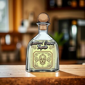 May include: A clear glass tequila bottle with a wooden stopper. The label reads "Emotional Support Tequila" and features a sugar skull design. The bottle sits on a wooden surface.