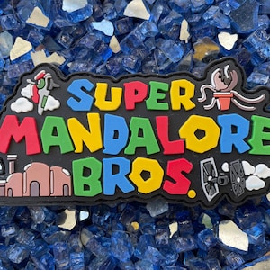May include: A black patch with the text "SUPER MANDALORE BROS." in colorful letters. The patch features various Star Wars characters and symbols, including a TIE fighter, a Mandalorian helmet, and a Tusken Raider.