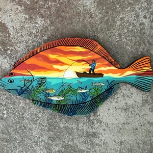 May include: A flat, fish-shaped wooden wall art piece featuring a vibrant sunset scene. The design depicts a person in a small boat fishing on a calm sea, with underwater details showing green seaweed and several small fish swimming below the surface.