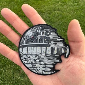 Könnte beinhalten: Ein schwarzer, grauer und weißer Death Star-Aufnäher mit detailliertem Design. Der Aufnäher ist rund mit einem fehlenden Abschnitt auf der rechten Seite. Der Aufnäher wird in einer Hand vor grünem Hintergrund gehalten.