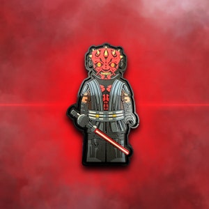 Może przedstawiać: Czerwono-czarna minifigurka Lego Dartha Maula, Lorda Sithów z Gwiezdnych Wojen. Trzyma czerwony miecz świetlny i ma rogatą maskę.