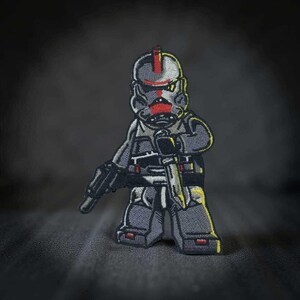 May include: Embroidered patch featuring a stylized soldier in gray and black, holding a blaster and a knife. The soldier's helmet has a red stripe. The patch has yellow accents and is set against a dark gray background.