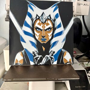 以下が含まれることがあります： スターウォーズのキャラクター、アソーカ・タノの印刷された画像のクローズアップ。青と白のヘッドウェアと黒と金の衣装を着ています。画像は正方形の木材に印刷されています。