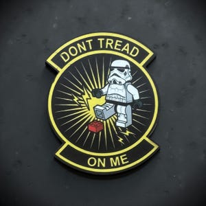 May include: A black and yellow patch with the text "DONT TREAD ON ME". The patch features a Stormtrooper figure stepping on a red Lego brick, with a yellow burst design in the background.