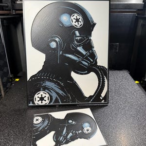 May include: Black and white artwork featuring a Star Wars TIE fighter pilot helmet. The artwork includes the Imperial symbol and a blue accent on the helmet. A smaller, similar artwork is also present.