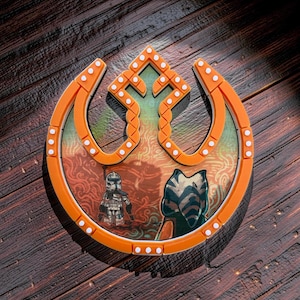 May include: Orange and white emblem with a Star Wars theme, featuring characters and a symbol. The emblem is set against a wooden background. The design includes a central image with two characters and a detailed orange border.