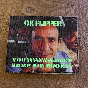 Peut inclure: Un patch noir et vert avec une image de Nicolas Cage et le texte "OK Flipper You Wanna Make Some Big Bucks?"