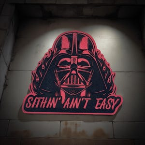 May include: A red and black graphic of Darth Vader's helmet with the text "SITHIN' AIN'T EASY". The design is set against a textured background, creating a striking image.