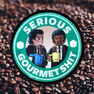 May include: A green and black circular patch with the text "Serious Gourmet Shit" in white. Two cartoon figures, one with black hair and one with an afro, are holding mugs of coffee.