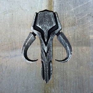 May include: A silver-colored Mandalorian symbol with a skull-like design and two curved horns. The symbol is made of a textured material and has a weathered look.