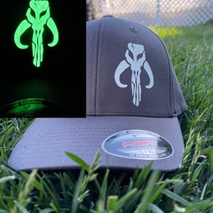 May include: A gray baseball cap with a white Mandalorian symbol on the front. The symbol glows green in the dark. The cap has a black Flexfit label on the back with the text "The Original Flexfit It Fits Your Life".
