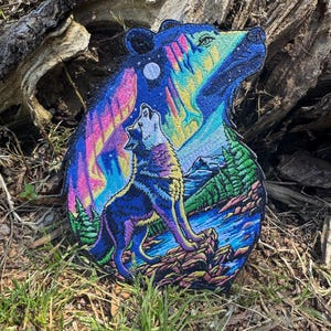 May include: A colorful embroidered patch featuring a bear-shaped design with a wolf howling at the moon. The patch showcases a vibrant night sky with aurora borealis, mountains, and a lake. The patch is placed on a bed of grass and wood.