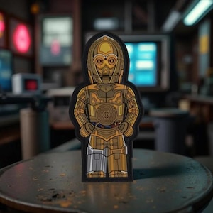 May include: A gold and bronze-colored robot character patch with a black border. The robot has a detailed design with visible mechanical components and is standing upright. The background is blurred, suggesting an indoor setting.