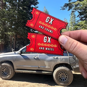 May include: Two red ticket-shaped stickers with the text "GX and Crawl" and "GX and Wheel" are held in front of a silver SUV. The stickers have a map-like design and the Lexus logo. The background shows trees and a blue sky.