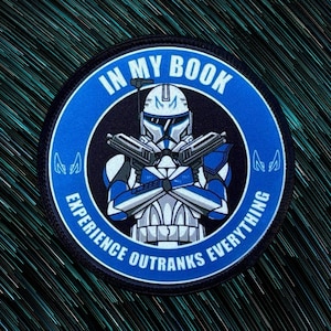 Peut inclure: Un patch circulaire bleu et noir avec une image de dessin animé d'un clone trooper Star Wars bleu et blanc tenant deux blasters. Le texte "In My Book Experience Outranks Everything" est imprimé autour de l'image.