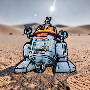 May include: Embroidered patch of a robot with a light blue body, orange and red accents, and black details. The robot has a unique design with arms and a head antenna. The patch is set against a desert backdrop.