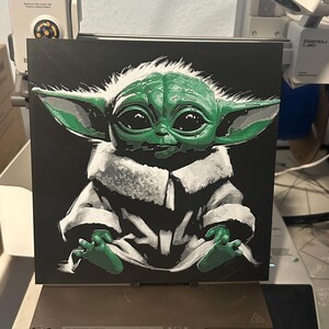 May include: A green and white 3D printed image of a character from the Star Wars franchise, known as "The Child" or "Baby Yoda", on a black background. The character is sitting with its hands in its lap.