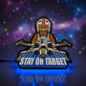 May include: A light-up sign featuring a cartoon pilot in an X-wing fighter. The pilot wears an orange flight suit and a white helmet. The sign has the text "STAY ON TARGET" in white on a blue background. The sign is illuminated with blue light.
