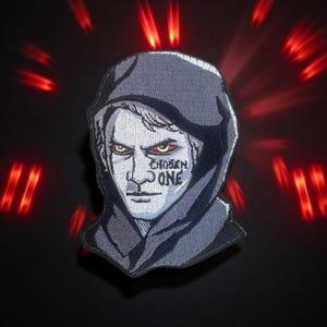 May include: Embroidered patch featuring a stern-faced figure in a hooded cloak. The figure has intense yellow eyes and the words "CHOSEN ONE" tattooed on their face. The patch is gray and black, with a red light burst effect in the background.