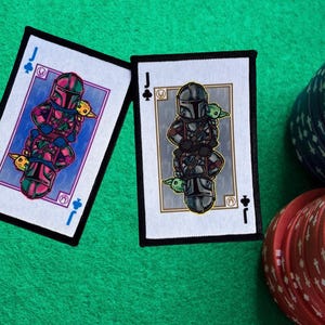 Może przedstawiać: Dwie karty do gry przedstawiające czarną i zieloną postać z hełmem i małym zielonym stworzeniem. Karty to obie walety, jedna to walet pik, a druga walet trefl.