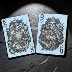 Può includere: Due carte da gioco, un re e una regina di cuori, su sfondo azzurro chiaro. Le carte presentano illustrazioni dettagliate di figure scheletriche in abiti reali. Il re tiene uno scettro e la regina tiene una rosa. Design unico.