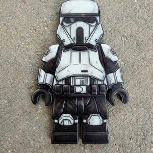 Könnte beinhalten: Eine flache, zweidimensionale Figur in Form eines Star Wars Trooper Minifiguren-Designs. Sie zeigt einen weißen Helm mit schwarzen Visierdetails, eine weiße Brustpanzerung und schwarze Gliedmaßen. Die Oberfläche hat ein verwittertes Finish.