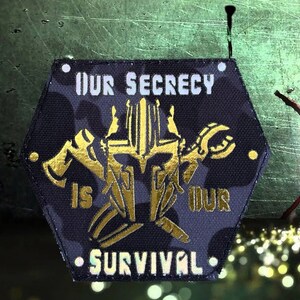 Könnte beinhalten: Ein schwarzer sechseckiger Patch mit goldener Stickerei. Der Patch zeigt einen Helm mit gekreuzten Äxten und einem Schraubenschlüssel. Der Text "OUR SECRECY IS OUR SURVIVAL" ist in Weiß gestickt.