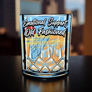 May include: A square-shaped rocks glass with the words "Emotional Support Old Fashioned" in white script. The glass features a colorful illustration of a cityscape in shades of blue, orange, and yellow. The glass has a black rim and a diamond pattern.