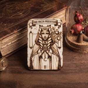 May include: A rectangular wooden plaque featuring a laser-engraved illustration of The High Priestess tarot card. The design depicts a figure with headgear, a crescent moon pendant, and a scroll, with the text THE HIGH PRIESTESS and the Roman numeral II.