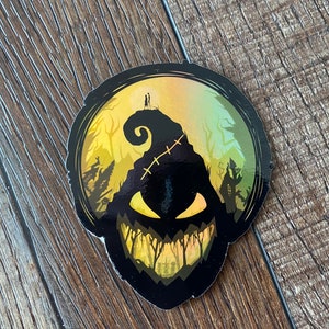 Op de afbeelding: Een zwarte en gele sticker met een silhouet van een griezelig, grijnzend personage met hechtingen op zijn hoofd. Het personage is geplaatst tegen een achtergrond van een bos met een huis in de verte.