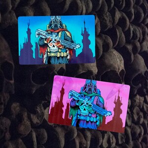 Op de afbeelding: Twee rechthoekige kaarten met een personage met een schedelvormig gezicht, helm en futuristisch wapen. De achtergrond van de bovenste kaart is blauw, die van de onderste kaart roze, beide met een silhouet van een stadsgezicht. Het personage draagt een donkere outfit met rode en groene accenten.