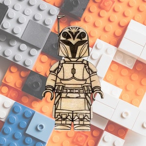 May include: A wooden figurine of a Mandalorian warrior, featuring detailed laser-etched armor and helmet design. The figure is set against a backdrop of colorful toy building blocks, including orange, white, gray, and blue blocks.