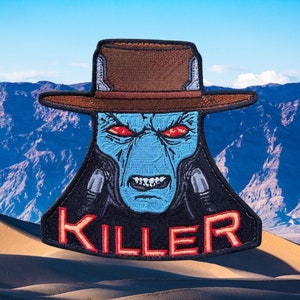 May include: A blue alien with red eyes wearing a brown cowboy hat and a black shirt with the word "KILLER" in red letters. The alien is against a background of blue mountains and sand dunes.